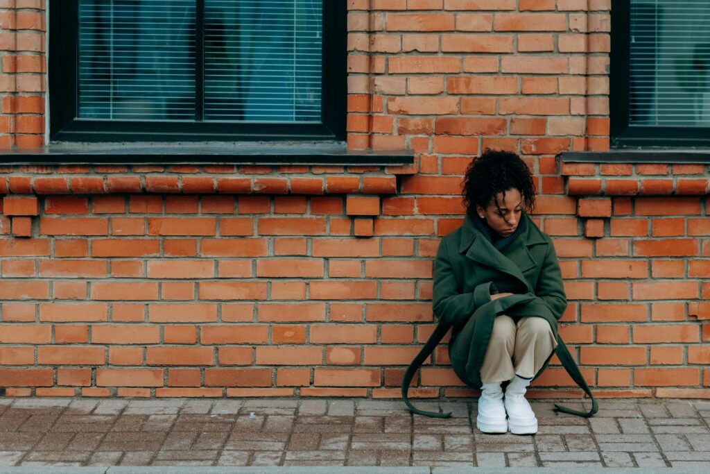 A woman in deep thought sits alone against a brick wall, conveying solitude.